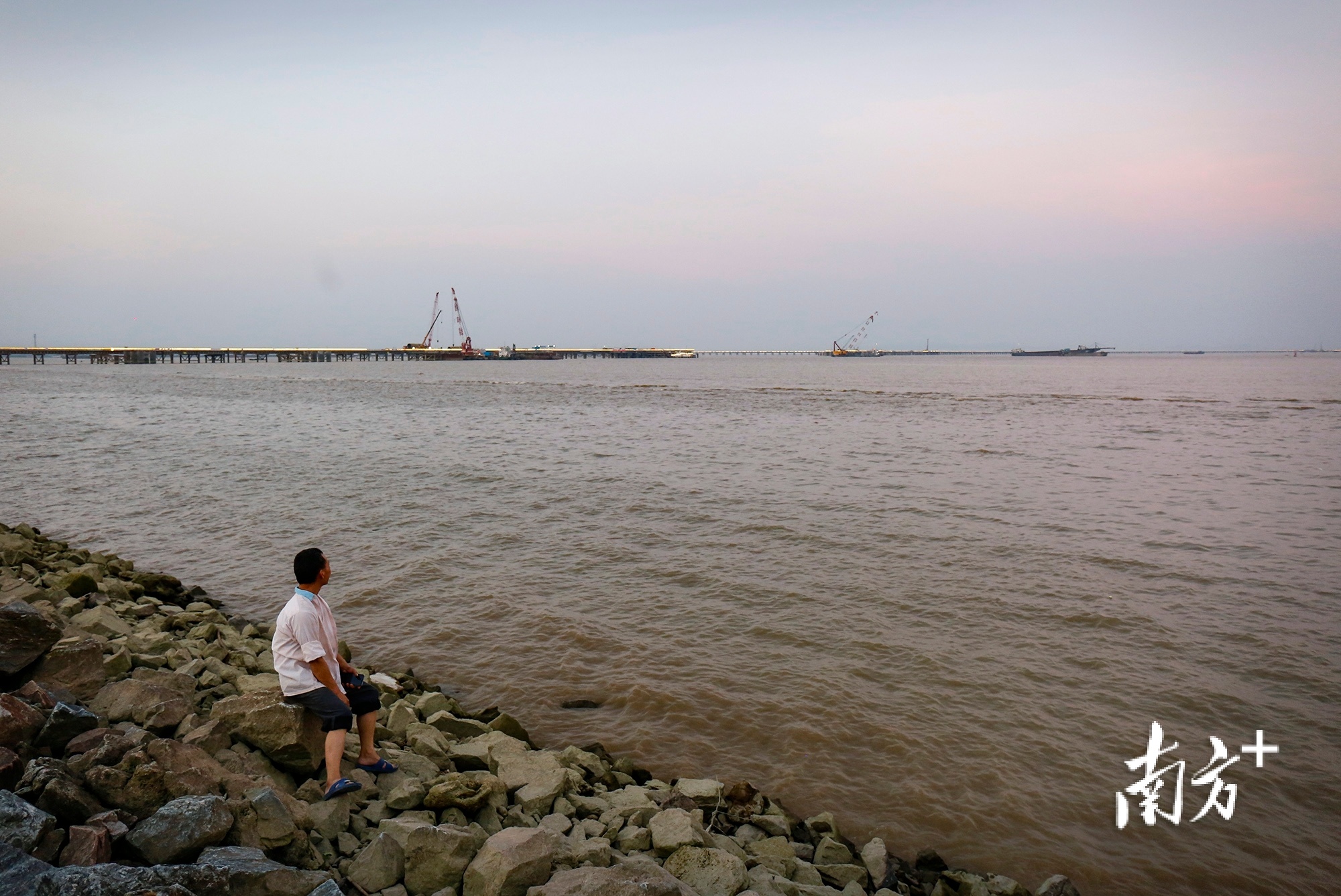 2019年7月16日，中山翠亨新区马鞍岛，在海边歇息的工人远眺建设中的深中通道。